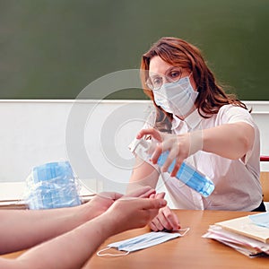 Woman schoolteacher treats hands with antiseptic. Female doctor in medical mask uses hand sanitizer sitting at a desk, copy space