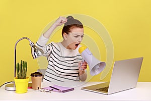 Woman screaming at megaphone, protesting, sitting on workplace and looking at laptop screen.