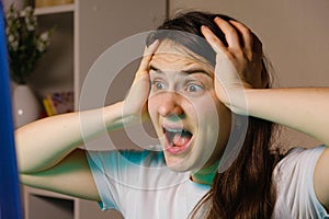 A woman screams and grabs her head with her hands while looking at a TV or computer screen.