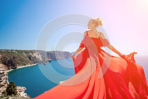 woman sea red dress. Blonde with long hair on a sunny seashore in a red flowing dress, back view, silk fabric waving in