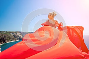 woman sea red dress. Blonde with long hair on a sunny seashore in a red flowing dress, back view, silk fabric waving in