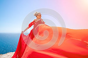 woman sea red dress. Blonde with long hair on a sunny seashore in a red flowing dress, back view, silk fabric waving in