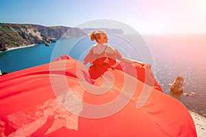 woman sea red dress. Blonde with long hair on a sunny seashore in a red flowing dress, back view, silk fabric waving in