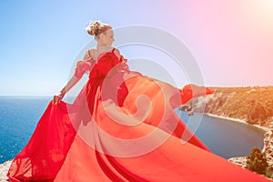woman sea red dress. Blonde with long hair on a sunny seashore in a red flowing dress, back view, silk fabric waving in