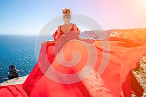 woman sea red dress. Blonde with long hair on a sunny seashore in a red flowing dress, back view, silk fabric waving in