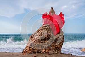 woman sea red dress. Woman with long hair on a sunny seashore in a red flowing dress, back view, silk fabric waving in