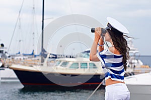 Woman in sea style clothes looks into the distance through binoculars on the background of yachts in the port
