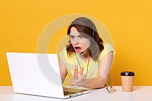 Woman seatting at table and looking at her laptop screen in consternation and horror, keeping mouth opened, wearinfcasual tshirt,