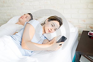 Woman Secretly Using Smartphone While Lying By Boyfriend On Bed