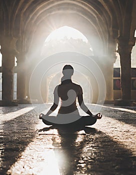 Woman seen from the back doing yoga pose against sun light in large hall