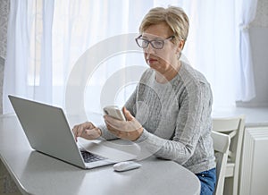 A woman senior work at a laptop with phone using modern technology in everyday life. online shopping