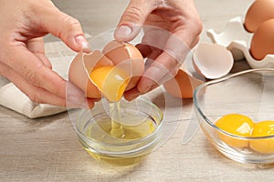 Woman separating egg yolk from white over glass bowl at wooden table, closeup