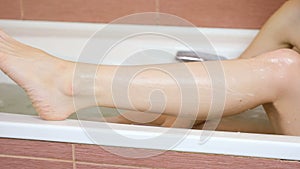 Woman shaving her leg in the bathroom with razor on the edge of the bath. Leg close-up.