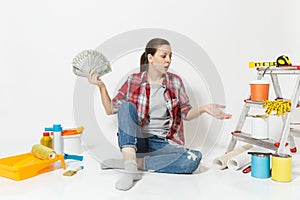 Woman in shock holds bundle of dollars, cash money, sits on floor with instruments for renovation apartment isolated on