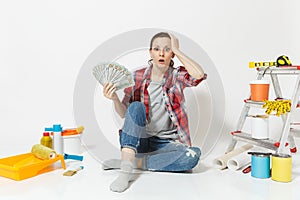 Woman in shock holds bundle of dollars, cash money, sits on floor with instruments for renovation apartment isolated on
