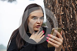 Woman is shocked and looks at phone screen in surprise when reading SMS or news, surprised by an unwanted call. outdoors