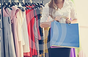 Woman shopping and holding shopping bag in women fashion clothing store with colorful women`s dresses on hangers in a retail shop