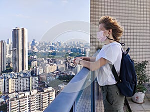 Woman with short blond hair in a medical mask with a backpack staying on the roof looking at the city scape