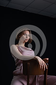 Woman with short hair posing in pink suit, sitting on chair on black background