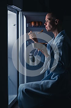 A woman with a short haircut eats near the refrigerator at night. Night appetite, diet concept. A bald thin woman satisfies her