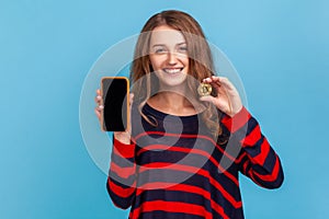 Woman showing golden bitcoin and smart phone with empty screen for advertisement, smiling to camera.