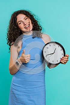 Woman showing time on wall office clock, ok, thumb up, approve, pointing finger at camera, your time