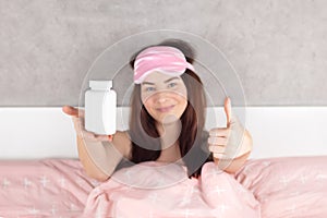 Woman shows a white jar for a good sleep, insomnia pills mock up. A beautiful girl in bed in pink sleep mask. She slept well and