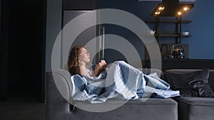 Woman sick with flu coughing and blowing her nose under blanket on couch at home. Young caucasian female suffering from