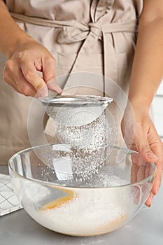 Woman sieving flour into bowl at light grey table, closeup. Cooking of delicious cake