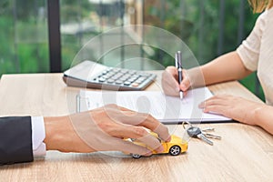 Woman signing car loan agreement contract with car key and calculator on wooden desk.