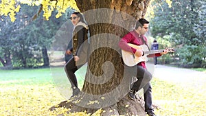 Woman singing and man playing guitar while leaning against tree in park
