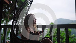 Woman sips tea in hanging chair overlooking mountainous tea plantation. Luxe balcony with green scenery, solo traveler