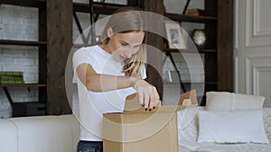 Woman sit on couch in living room open cardboard box shopping online at home, happy female unpack parcel order purchase