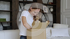 Woman sit on couch in living room open cardboard box shopping online at home, happy female unpack parcel order purchase