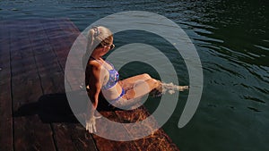 Woman sit on a sunbed in sunglasses and swimming suit. Girl rest on a flood wood underwater pier. The pavement is