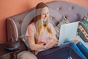 A woman sits on a bed and works on a laptop. Aroma diffuser next to the bed