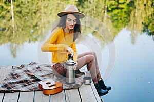 Woman sits on a bridge on a lake with an autumn landscape and drinks hot tea