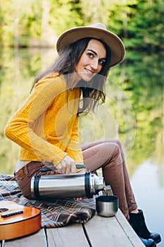 Woman sits on a bridge on a lake with an autumn landscape and drinks hot tea