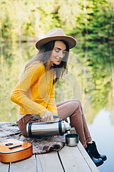 Woman sits on a bridge on a lake with an autumn landscape and drinks hot tea