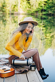 Woman sits on a bridge on a lake with an autumn landscape and drinks hot tea