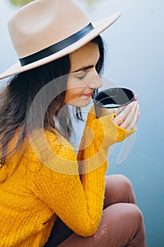 Woman sits on a bridge on a lake with an autumn landscape and drinks hot tea