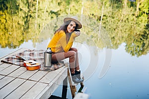 Woman sits on a bridge on a lake with an autumn landscape and drinks hot tea