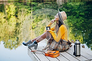 Woman sits on a bridge on a lake with an autumn landscape and drinks hot tea