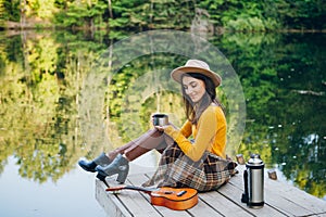 Woman sits on a bridge on a lake with an autumn landscape and drinks hot tea