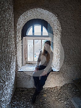 A woman sits by a broken window in an old abandoned dark building. Journey through ancient ruins