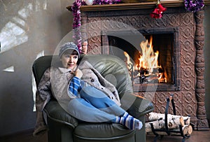 A woman sits in a chair near a burning fireplace
