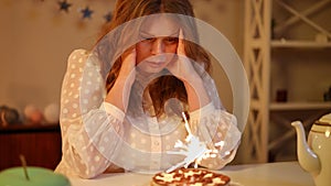 A woman sits, with clasp her head in her hands, at a table with a burning sparkler in a birthday cake. A woman carefully