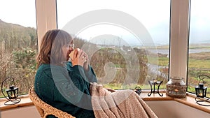 Woman sits in a cozy wicker chair with a plaid, on a light closed veranda in the mountains, drinks hot tea and looks out window,