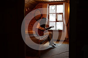 Woman sits on edge of bed in daylit wooden room in country house, play guitar with head bowed alone.