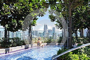 A woman sits on the edge of a pool surrounded by trees and plants with views of the city skyscrapers in the background